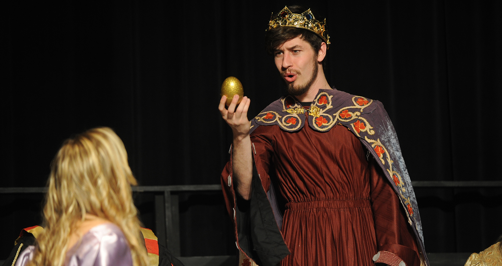 A student dressed as a king offers a golden egg to one of his servants